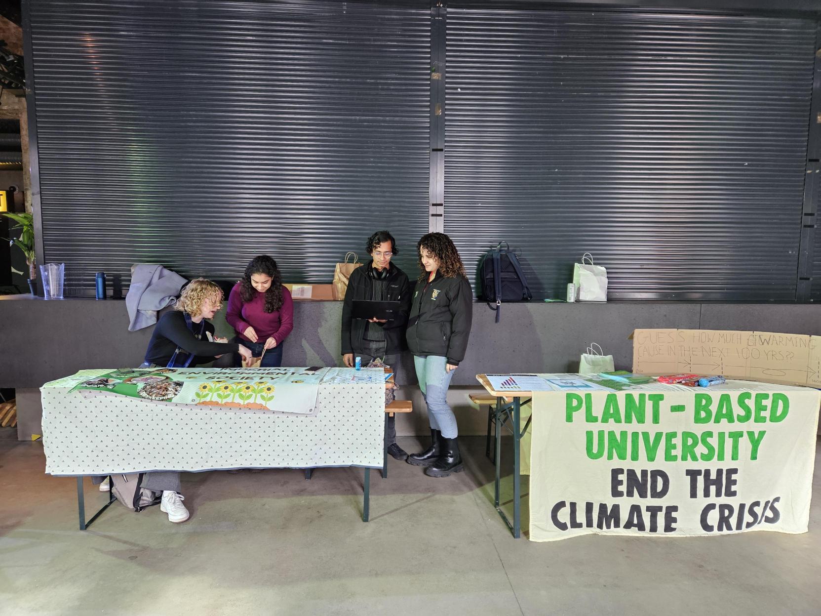 2 Tables, one has a banner over it which reads "Plant based university. End the climate crisis." There are 4 students behind the tables chatting.